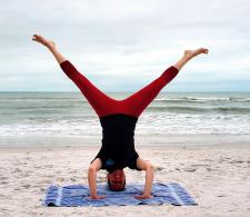 "a woman does a headstand with her legs open in a v on a beach with the ocean behind her"