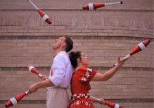 "two people in costume stand back to back and juggle bowling pins with each other"