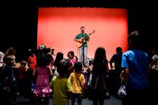 "a man, steve weeks, stands on a stage playing the guitar in front of kids and parents"