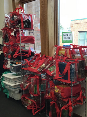 clear bags with red handles on metal shelves by large windows. the bags all have crafts and robotic kits inside.